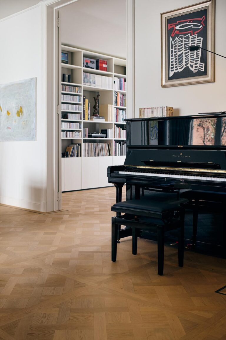 Eiken parketvloer met geometrisch Versailles patroon in een elegante woonkamer met zwarte vleugelpiano en ingebouwde bibliotheekkast